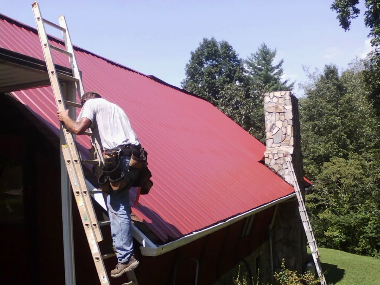 Roofer on a ladder beside a red metal roof and stone chimney — Soffit & Fascia Repair by Roofing Brothers in Aldie